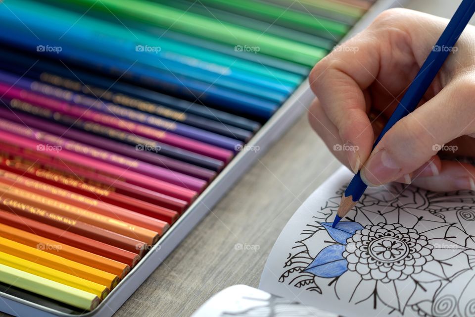 A portrait of a hand coloring in a drawing in a book. the other color pencils are still in the box in front of the book.