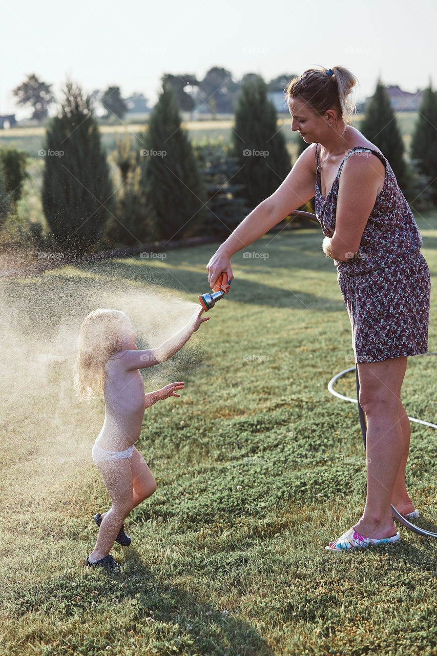 Little cute adorable girl enjoying a cool water sprayed by her mother during hot summer day in backyard. Candid people, real moments, authentic situations