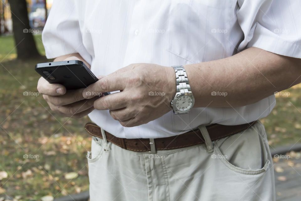 An elderly man walks alone in the park in the summer. A modern pensioner, businessman in a white shirt and trousers takes pictures with a camera in a mobile phone.