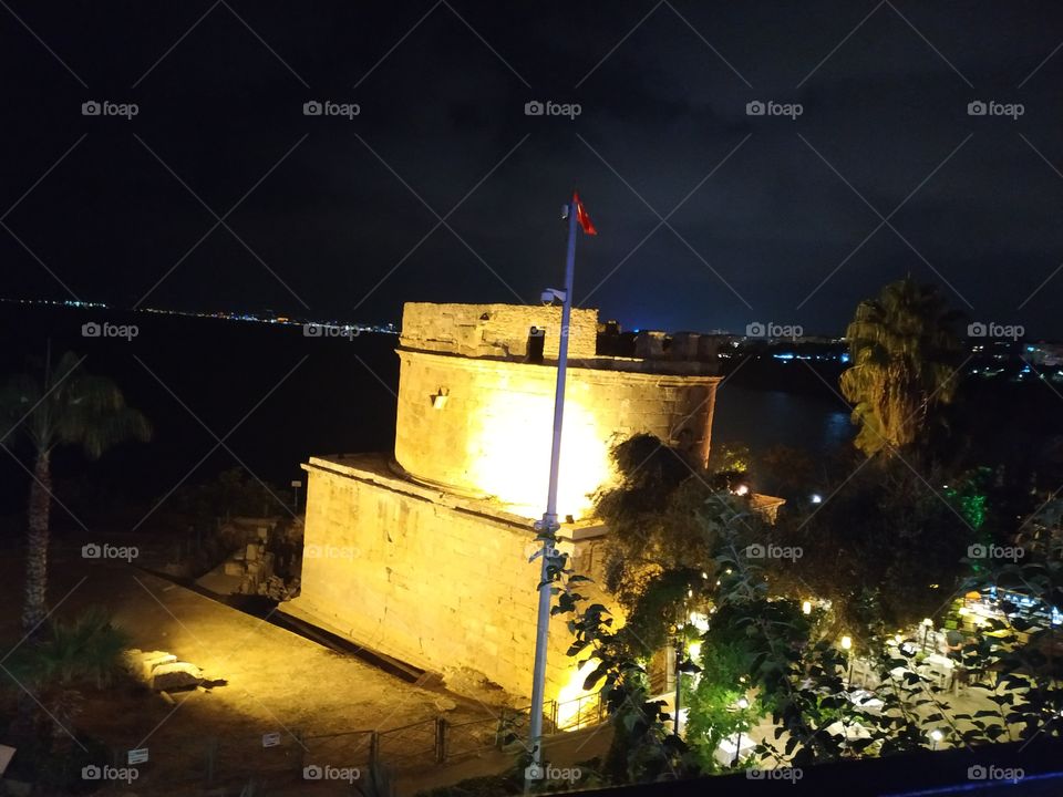 A beautiful old turkish iluminated building by the sea with seashore with iluminated city in background. Night view.