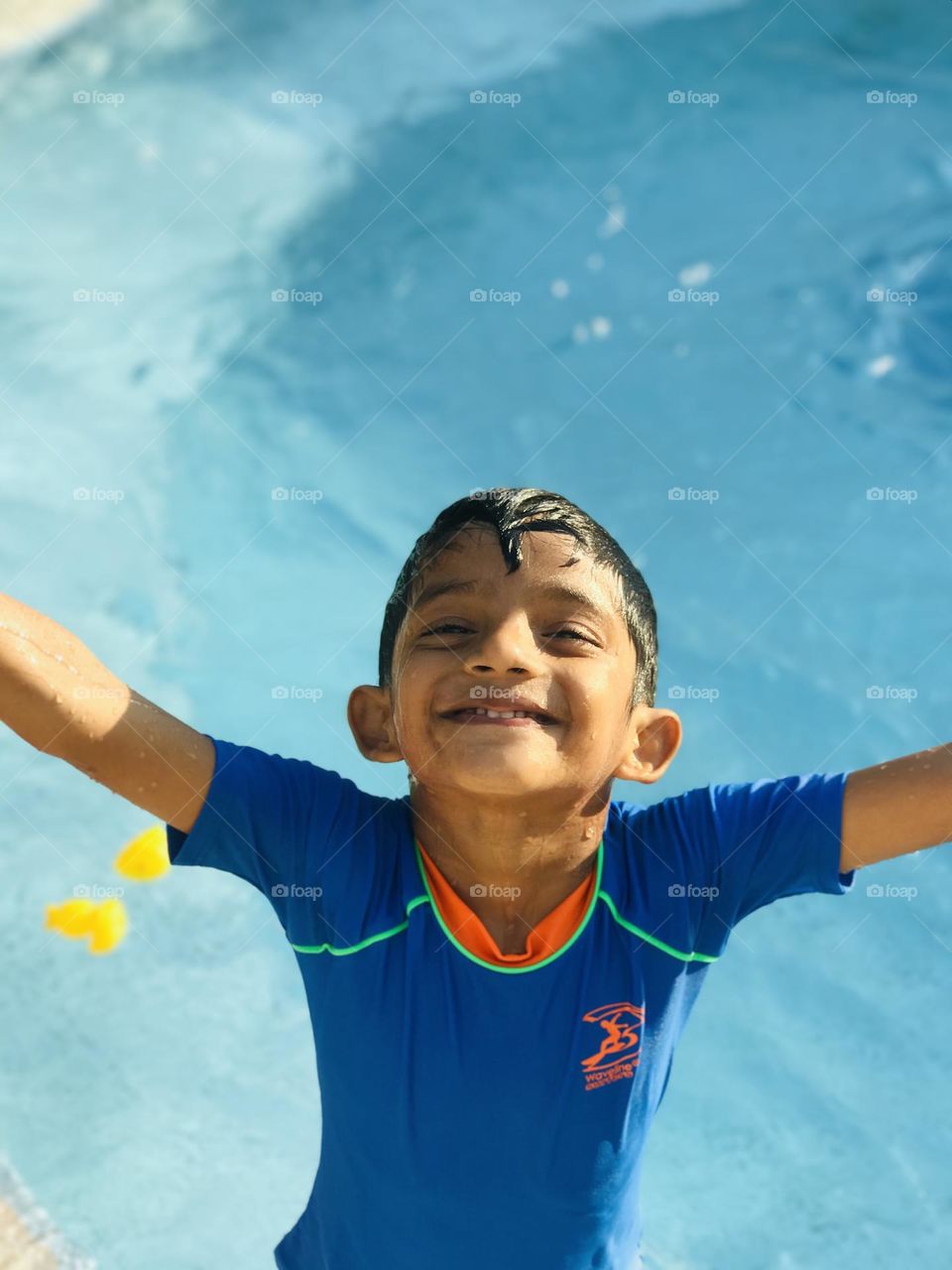 A 6 year old  boy enjoying a lot at swimming pool with cheerful face in summer and he is wearing blue colour swimsuit and he looks upside towards the sky by spreading hands both sides 😊