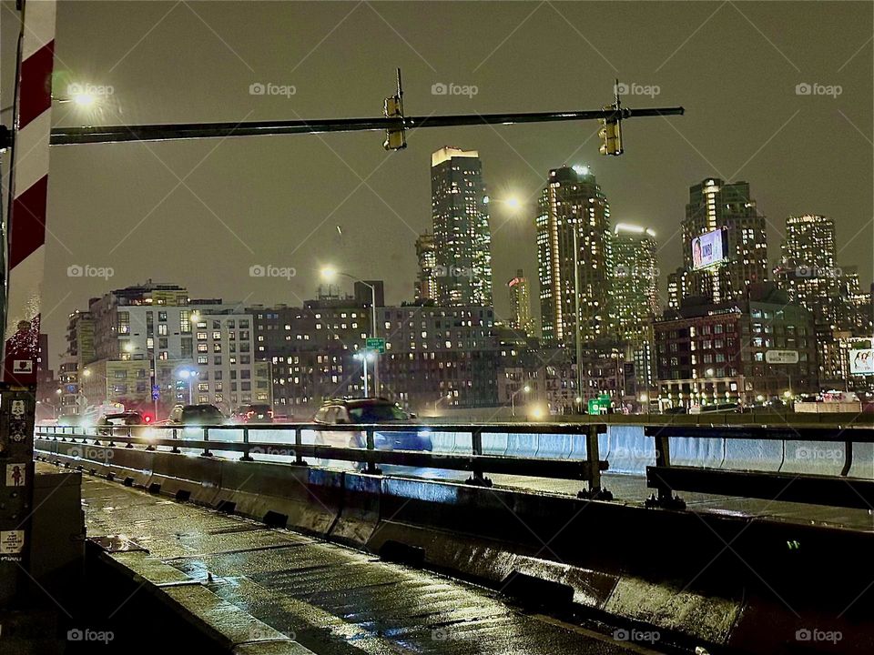 This nighttime impression comes from the pedestrian lane of the “Pulaski Bridge” at “Newtown Creek” that connects “Greenpoint”, Brooklyn and LIC, Queens. The cars with their bright headlights are speeding by incessantly. 2023. Hypnotic Productions