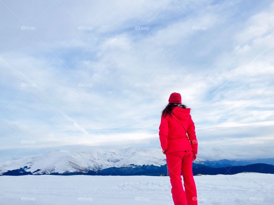 Back of woman hiker wearing red winter clothes surrounded by mountains covered in snow