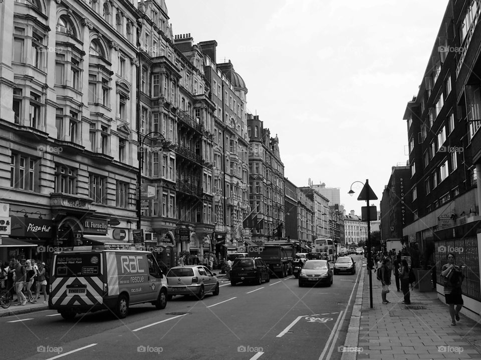 People walking and vehicles moving and parked on a busy ornate building lined buildings with shops on a busy street in London on a sunny summer day. 