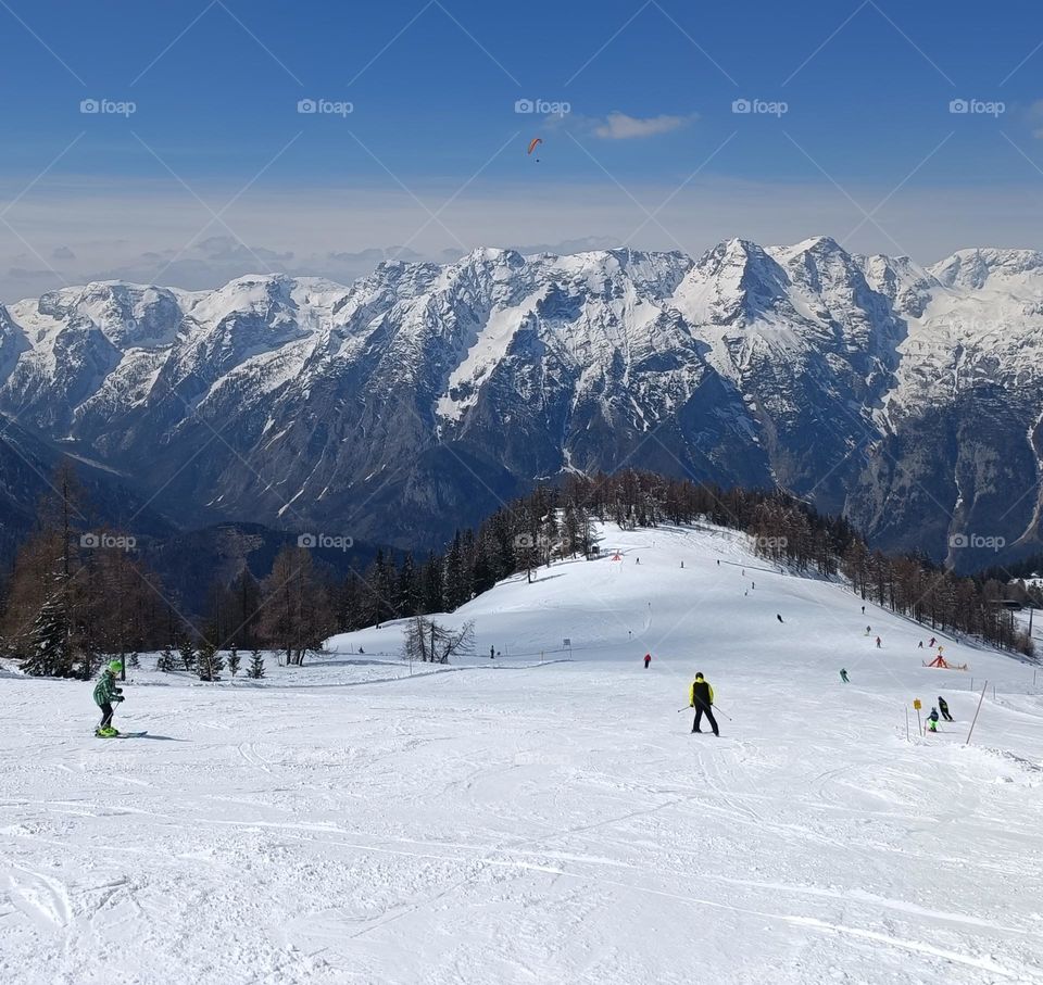 Landscape photography of a alpine ski piste with skiing people and a snow capped mountainscape at the background under a blue sky