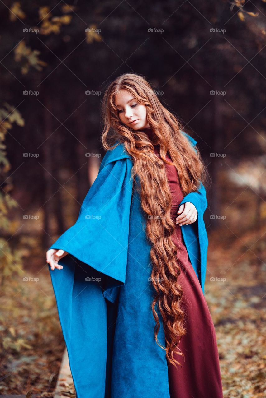 Beautiful girl with long red hair in blue coat. Woman staying in the forest