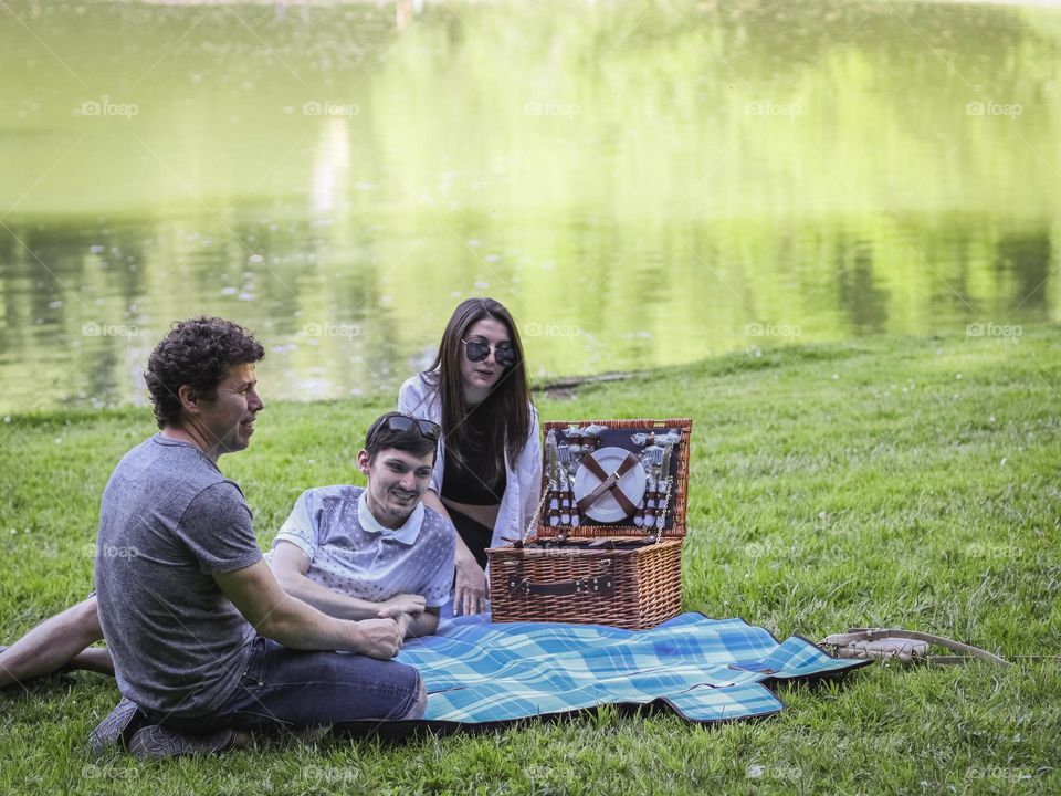 Three young caucasian people sit on a blanket with a wicker basket in a meadow near the lake and communicate with each other with happy smiles, close-up side view.