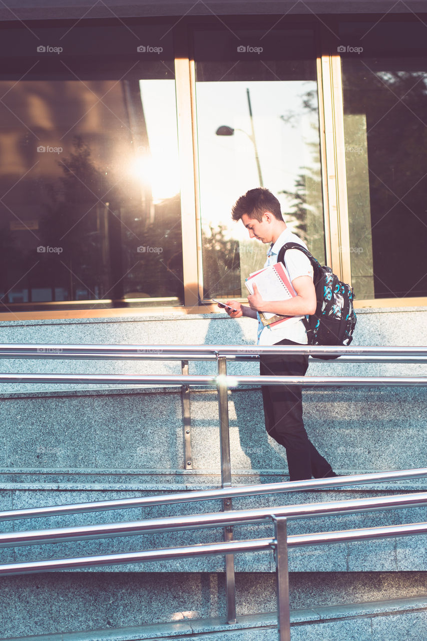 Student using mobile phone holding a notebook and carrying a backpack walking at the front of university building. Young boy wearing blue shirt and dark jeans