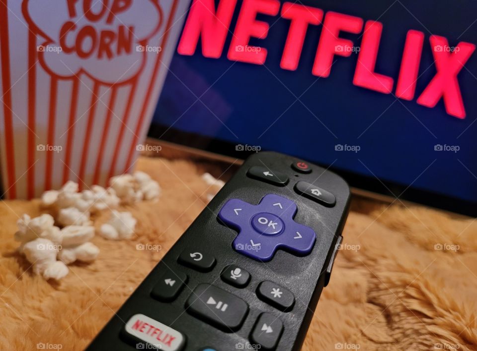 Aiming the remote control at television screen to choose favorite netflix channel to binge watch TV shows or movies for a fun and cozy night in with a popcorn snack on a soft blanket in bed