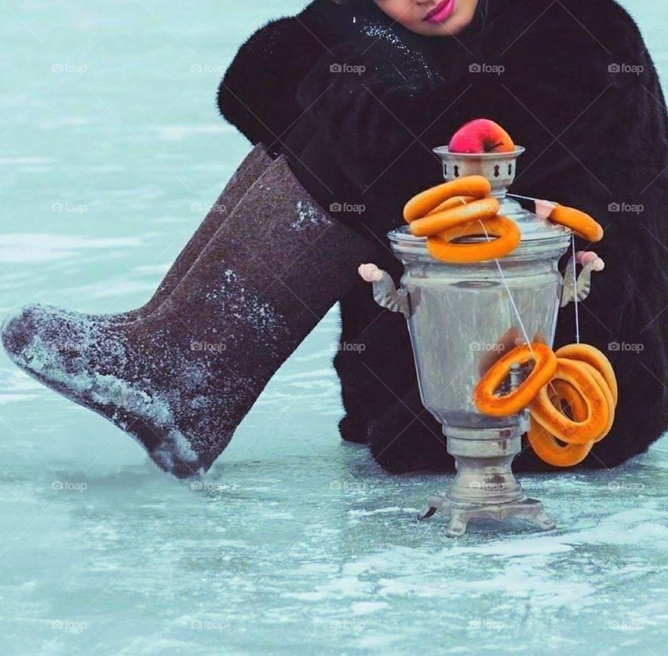 A girl sits on the ice in a fur coat and felt boots. There is a samovar on the ice with bagels hanging on it