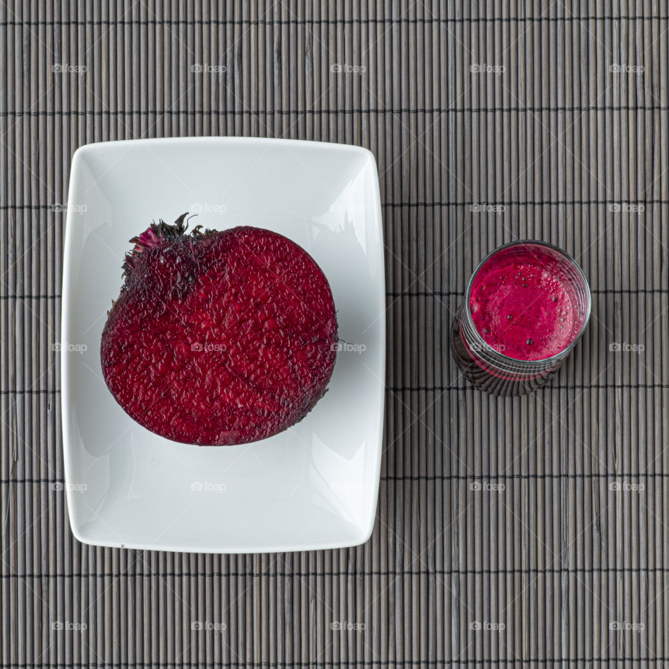 View from above half beetroot inside rectangular white dish next to transparent glass with beetjuice in a grey striped background pattern
