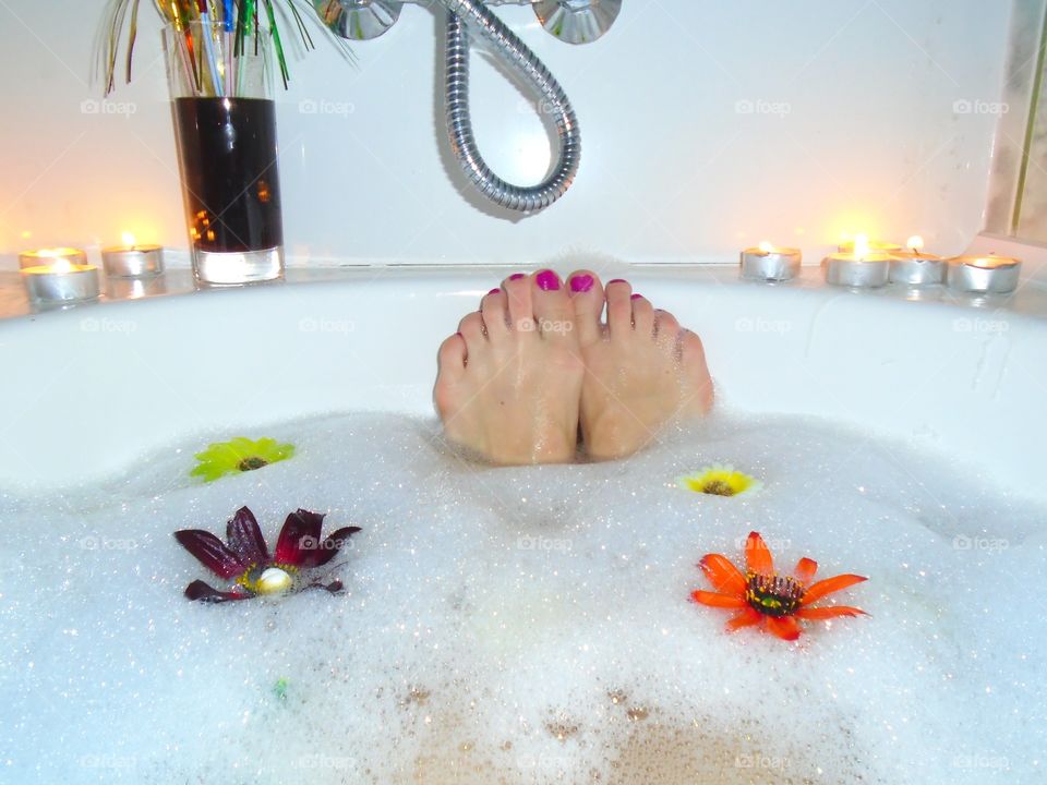 Woman's feet with red nail polish in bubble bath with candles and flowers