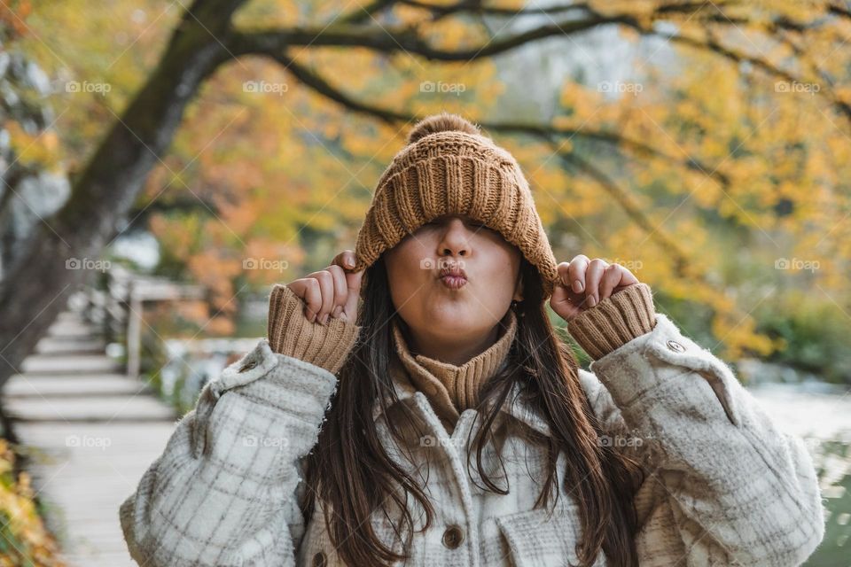 Portrait of happy girl standing on wooden footpath in forest in autumn. Young woman pulling knitted hat down over her eyes, puckering up toward camera.