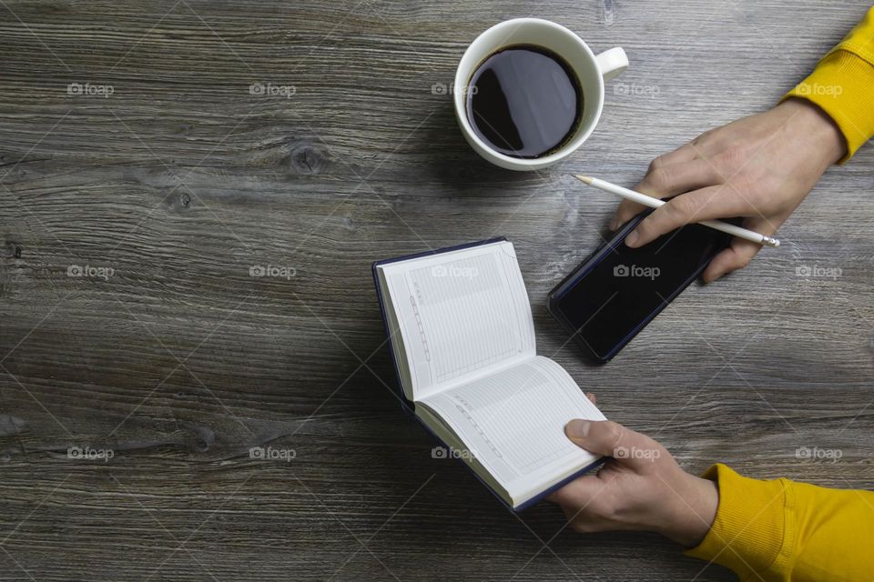A man drinks coffee in the morning, works on the phone and makes notes in pencil in a notebook. Men's hands hold a white cup of coffee and a mobile phone in a yellow jacket. The hands lie on a gray wood background.