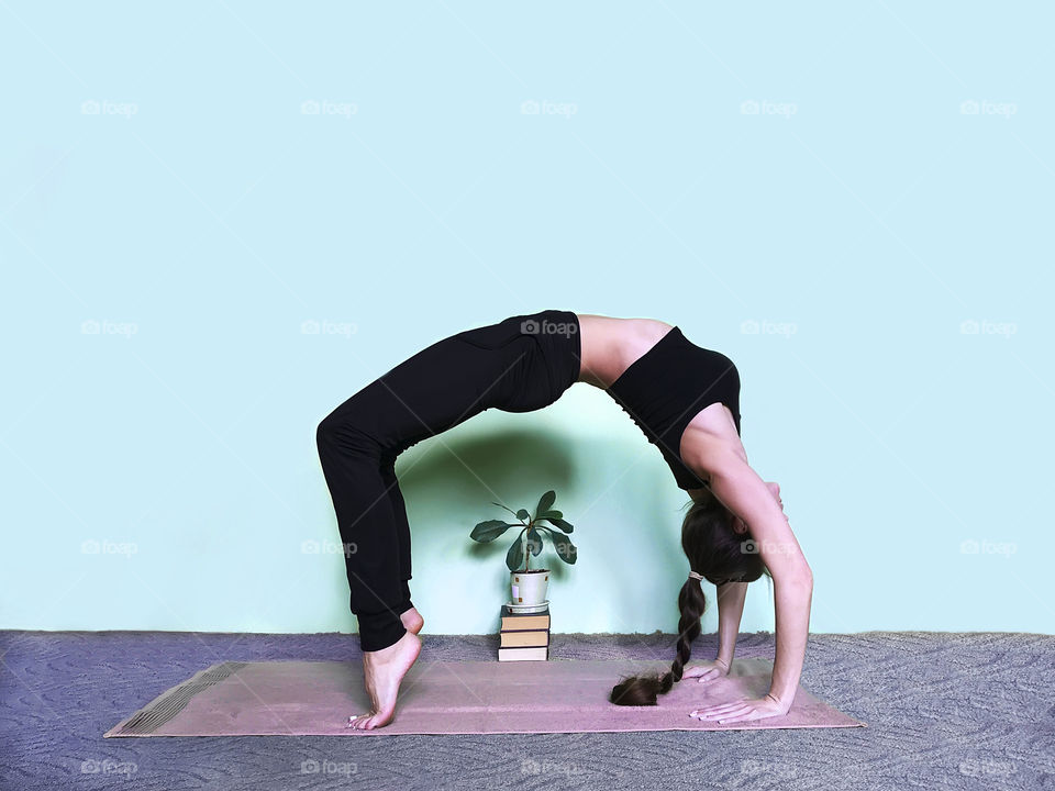 Young woman with long hair doing yoga at home nearby books and house plant on blue background 