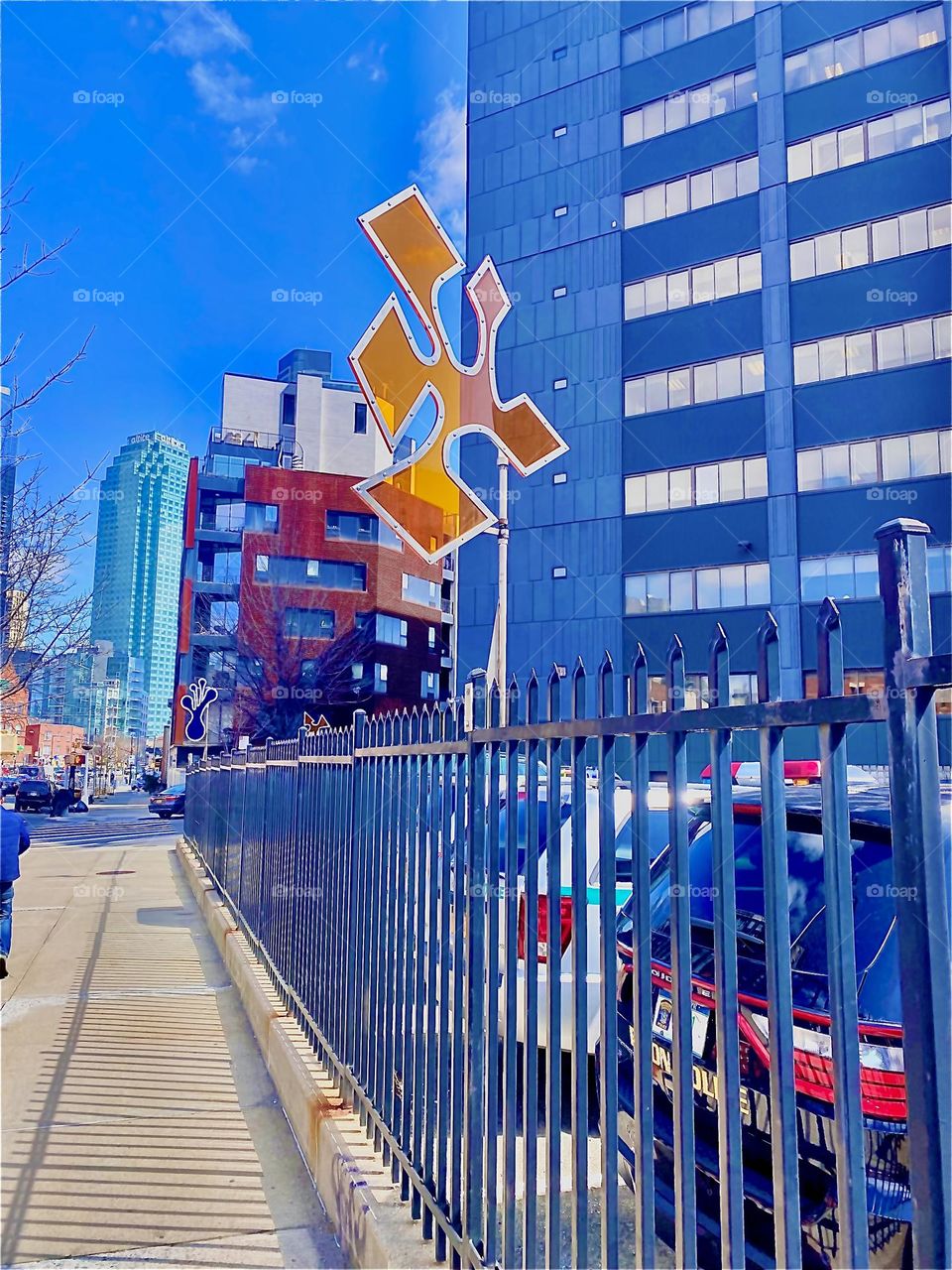 A semi transparent bright orange shape reminiscent of the pieces to a jigsaw puzzle enclosed by a solid border sits atop a pole above a metal fence attached so it spins whenever the wind blows at it. 2022. Hypnotic Productions