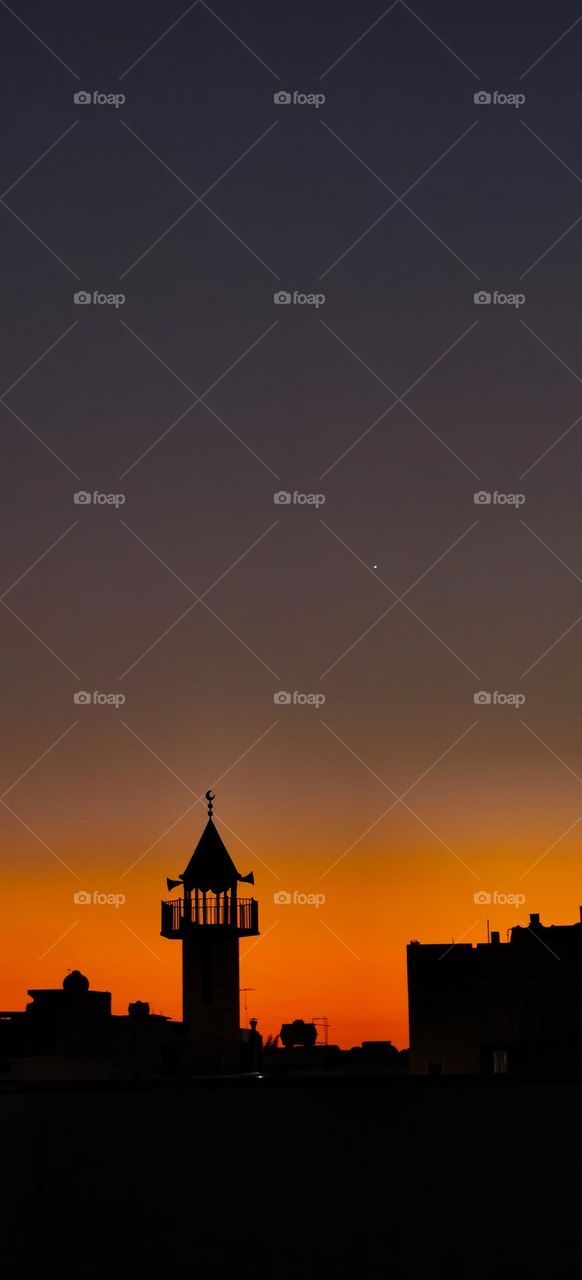 Orange sky at sunset with silhouette of mosque and houses with bright star shining in the dark sky