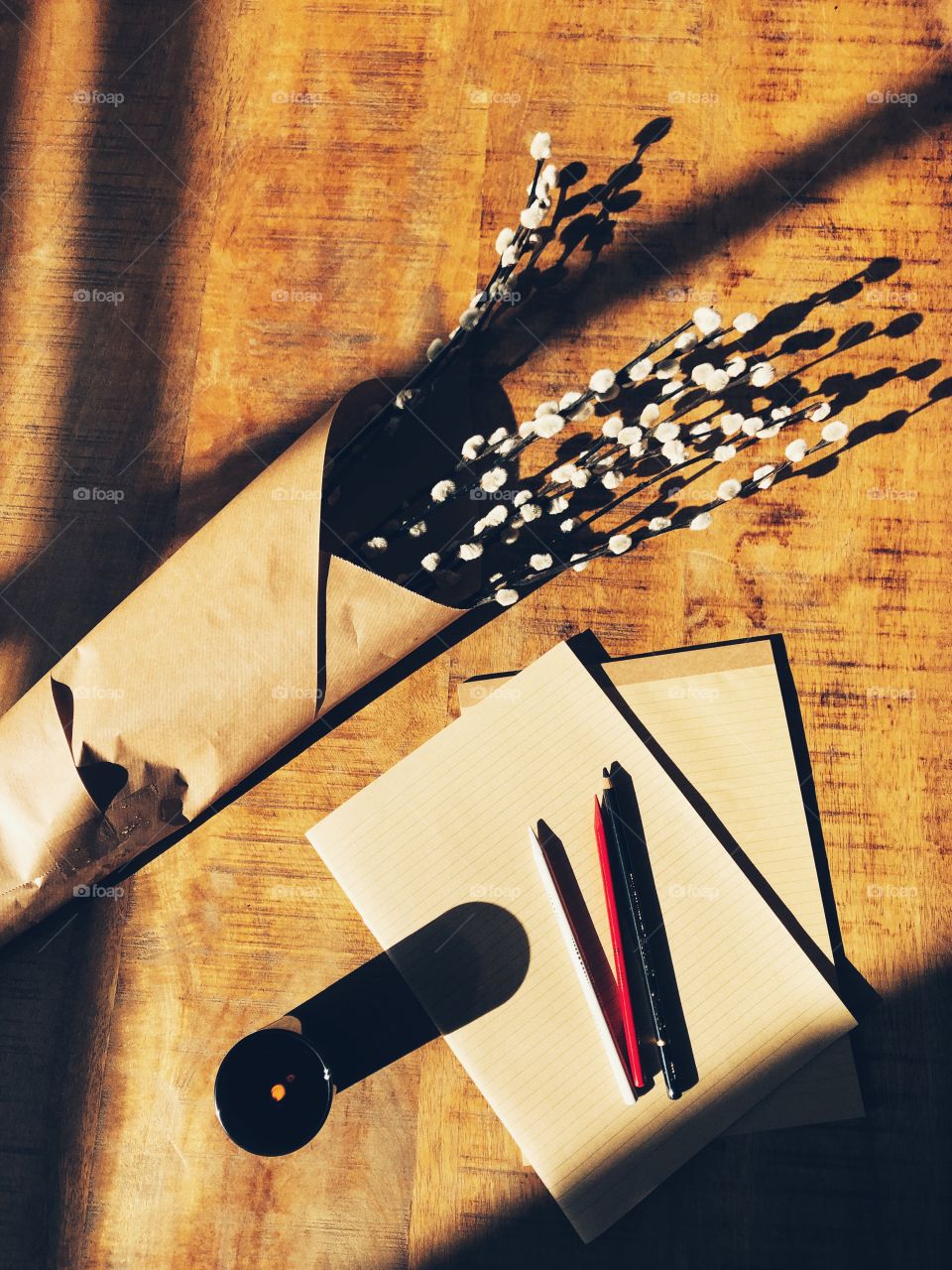 Bunched willow wrapped in rustic brown paper with brown paper notebooks, colored pencils and black candle in shafts of bright afternoon sunlight glow