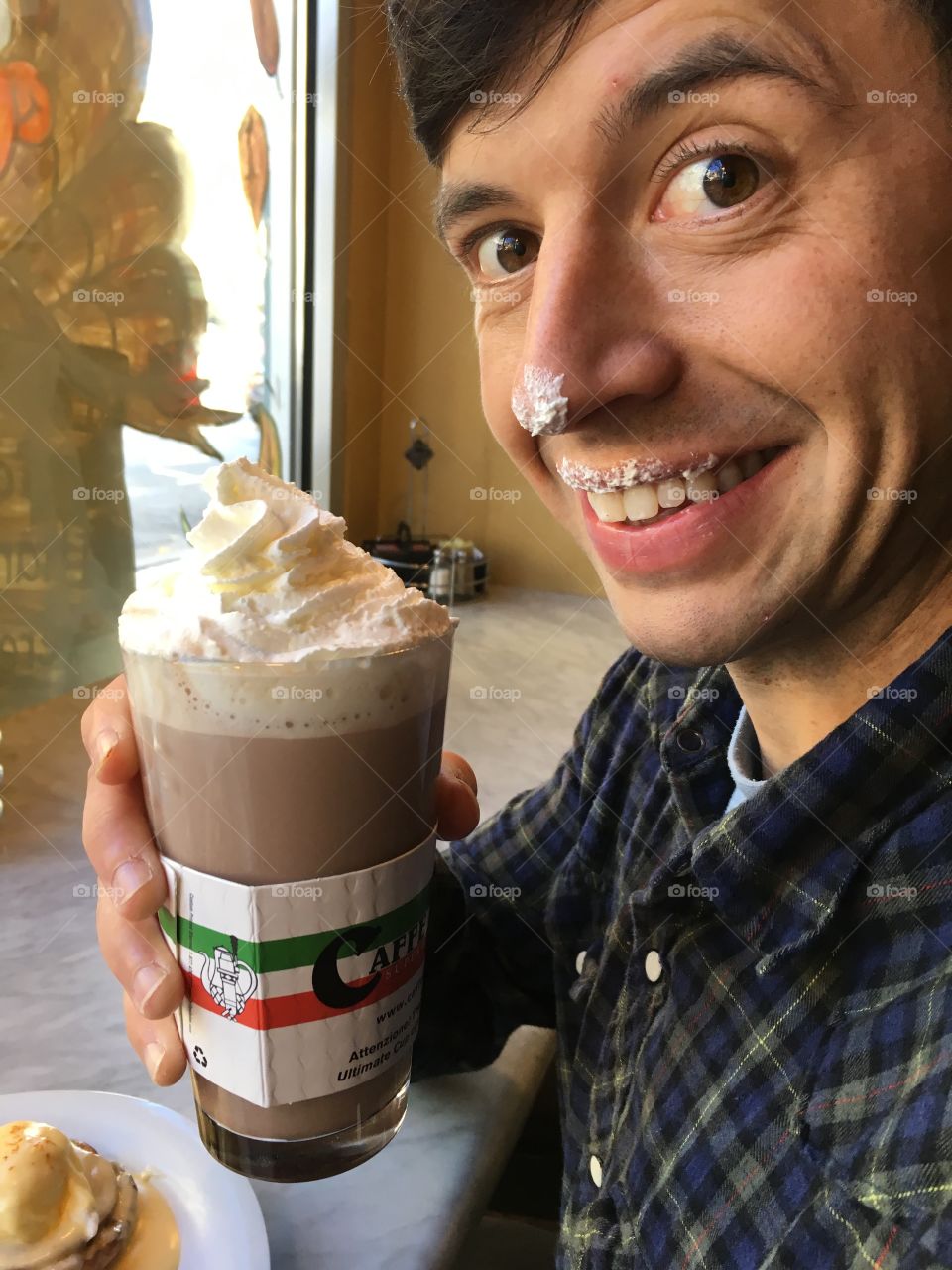 Man holding a whipped cream topped coffee beverage who is smiling with whipped cream on his nose and face