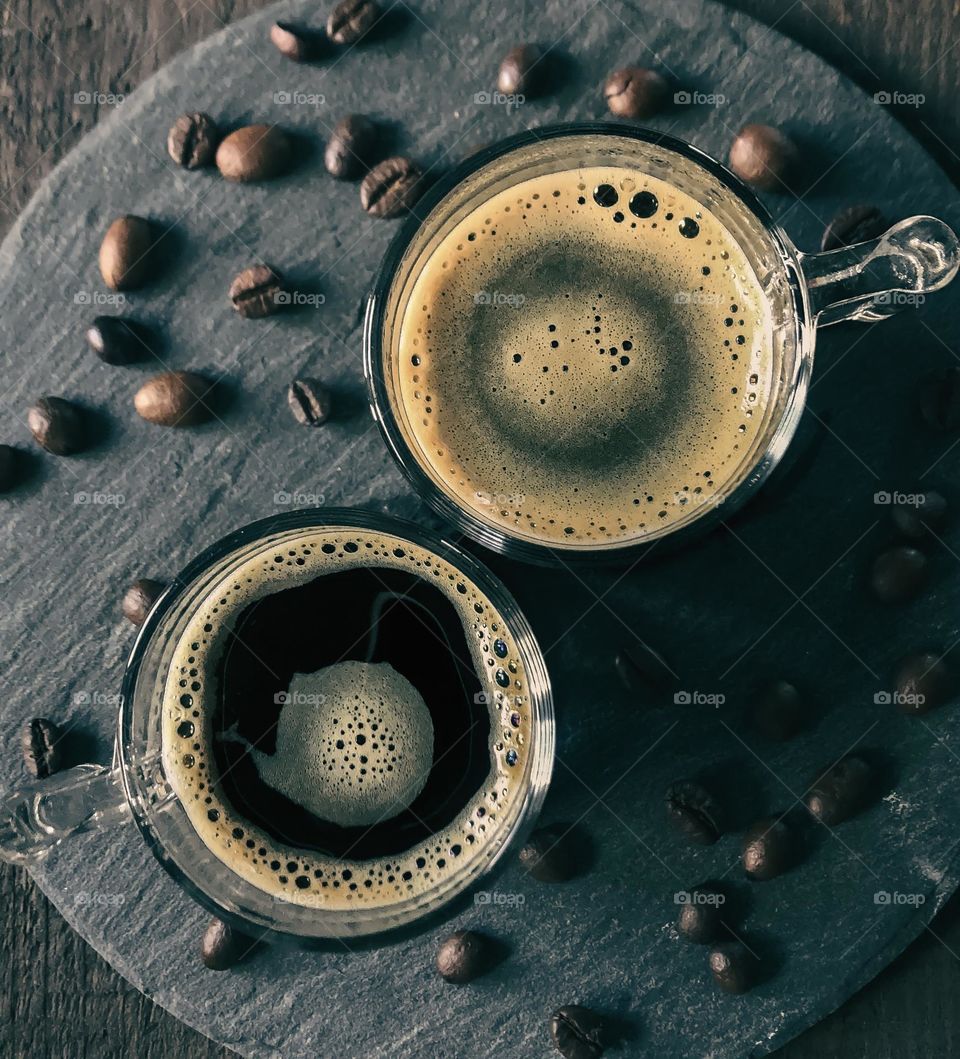 From above view of two black coffees in small glass mugs, on slate & surrounded by coffee beans