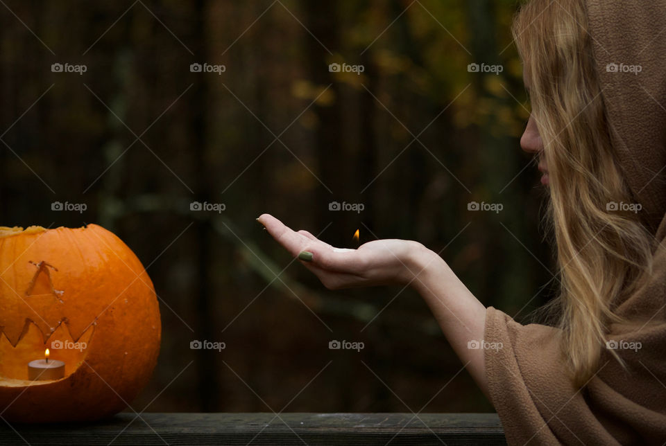 Igniting Life into a Pumpkin; Woman in Hooded Cloak holding a flame in the palm of her hand, extending it towards a cut pumpkin. 