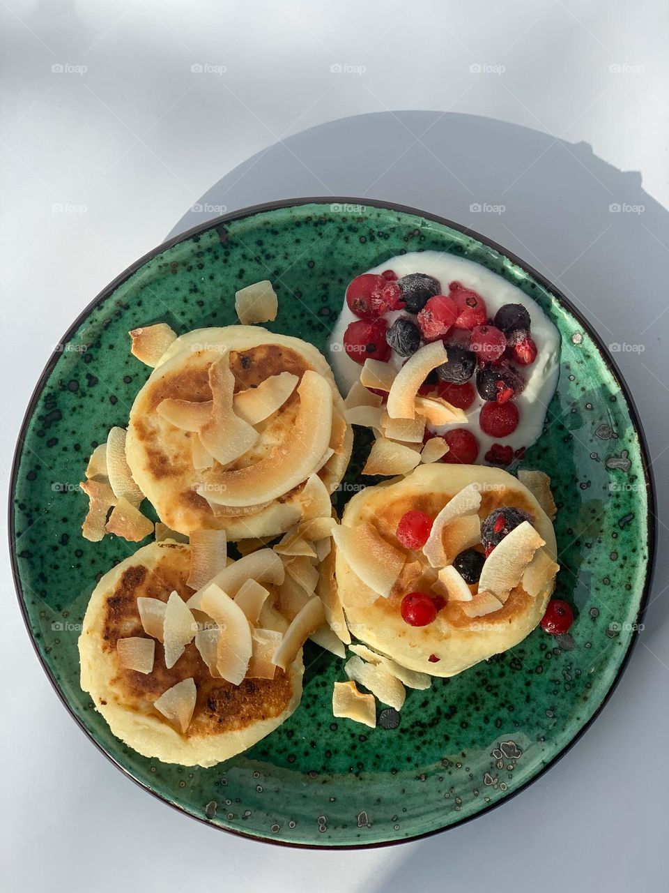 Delicious cottage cheese pancakes with yogurt, frozen berries and coconut chips flakes for breakfast. Healthy breakfast, nice fancy green plate in the morning sun