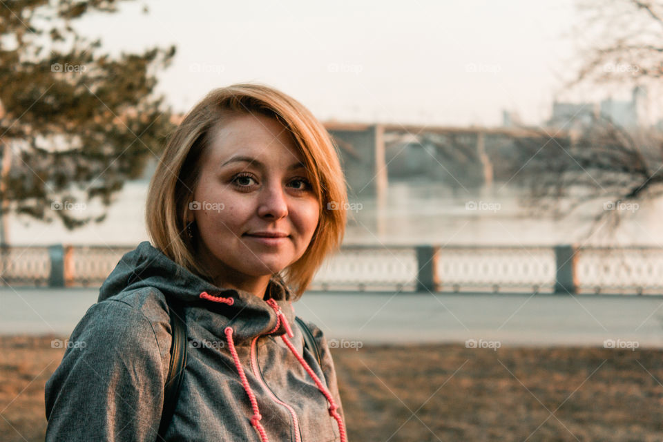 Beautiful young woman in the sunny spring park near bridge is looking to the camera at evening while sunset.