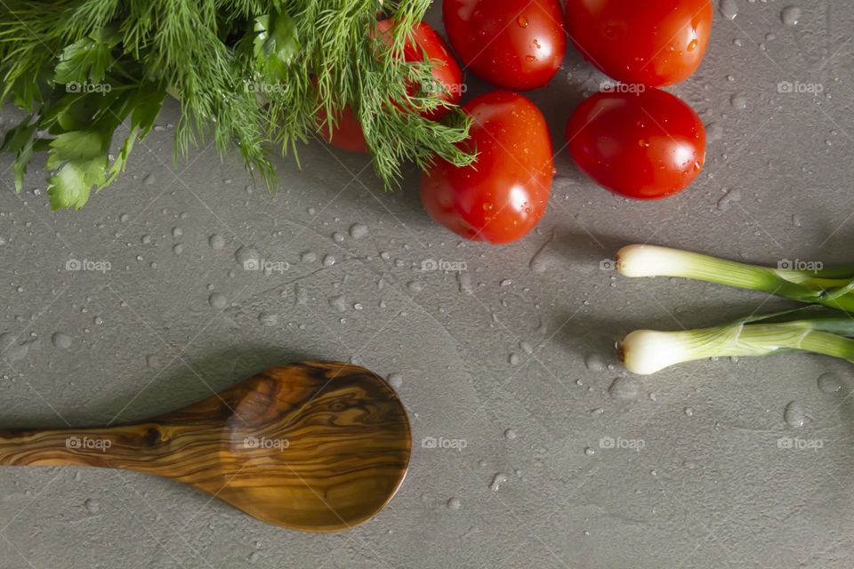 Fresh, bright, juicy vegetables, a bamboo spoon and herbs for salads and other vegetable dishes, are on a gray, kitchen worktop.