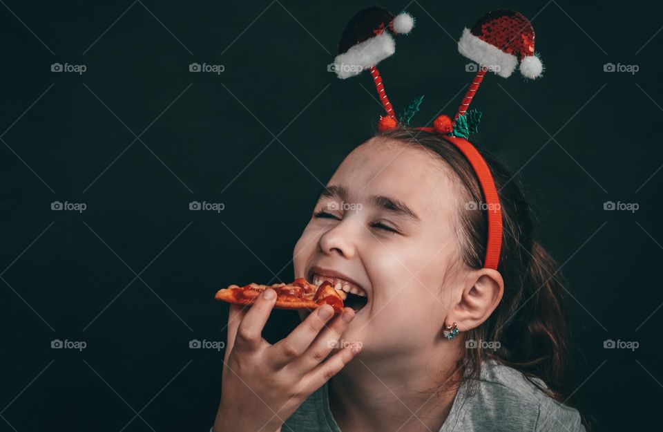 Portrait of one beautiful little caucasian girl in a headband from two santa claus hats with a happy smile and closed eyes bites off a fragrant tasty piece of homemade pizza on a black background, close-up side view. Happiness moment concept.