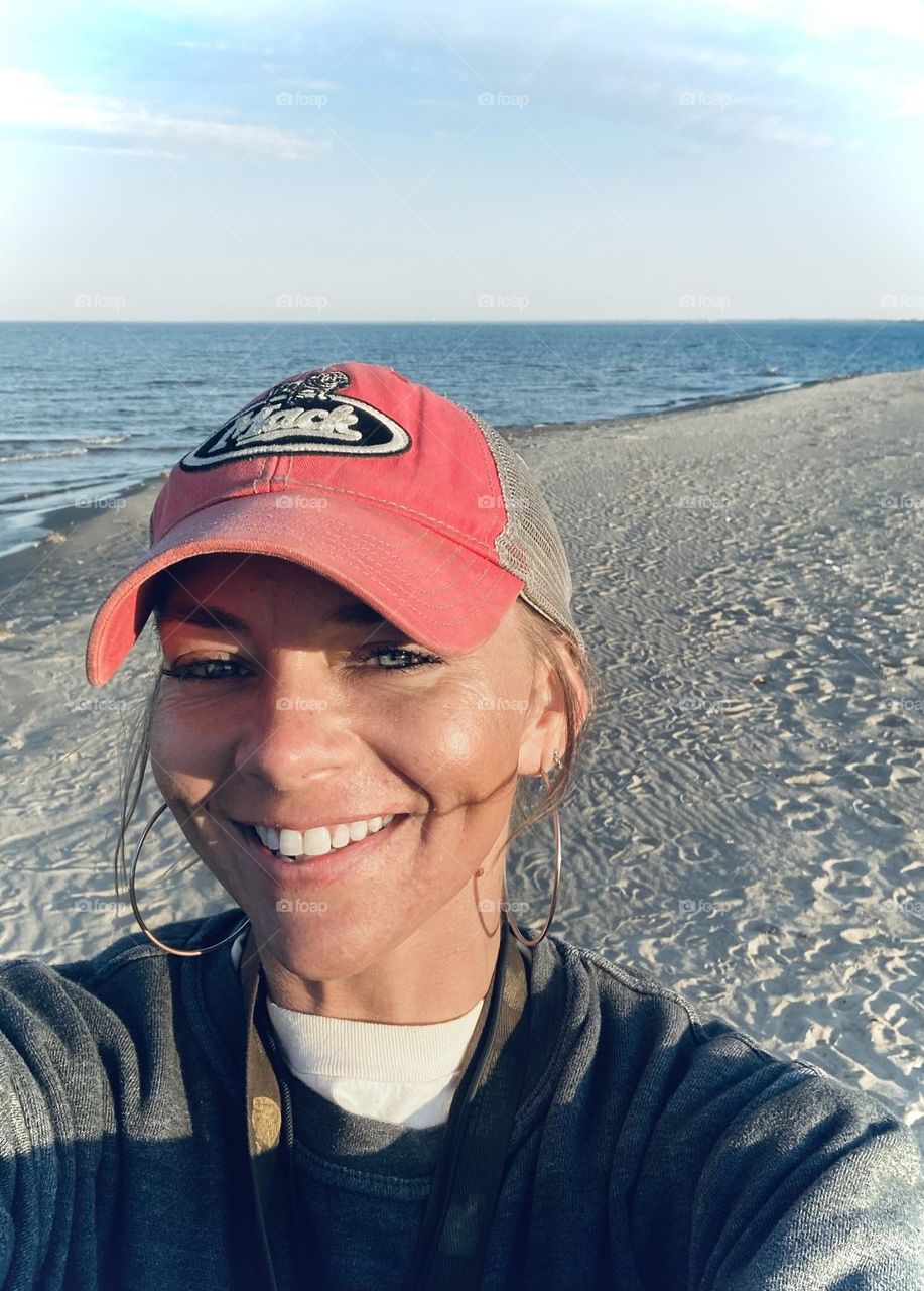 Smiling woman on Lake Eerie sandy shore wearing a red trucker hat, hoop earrings and and big smile