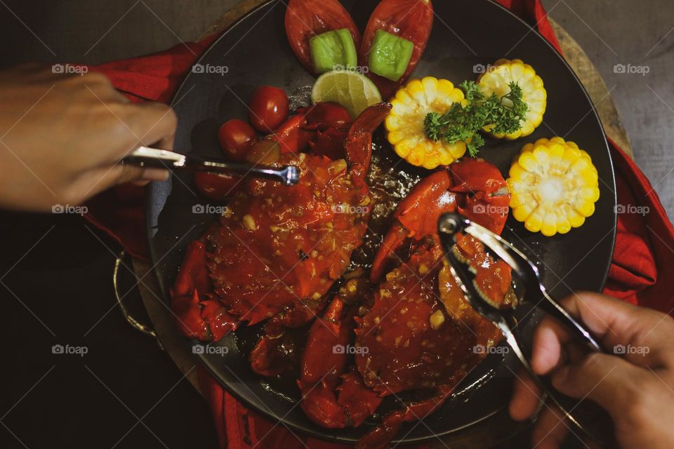 Crab flavored in creamy garlic sauce served with corn, tomatoes, lemon wedges and cherries on a black plate set on a red cloth set in a wooden setting. Asian flavor crab.