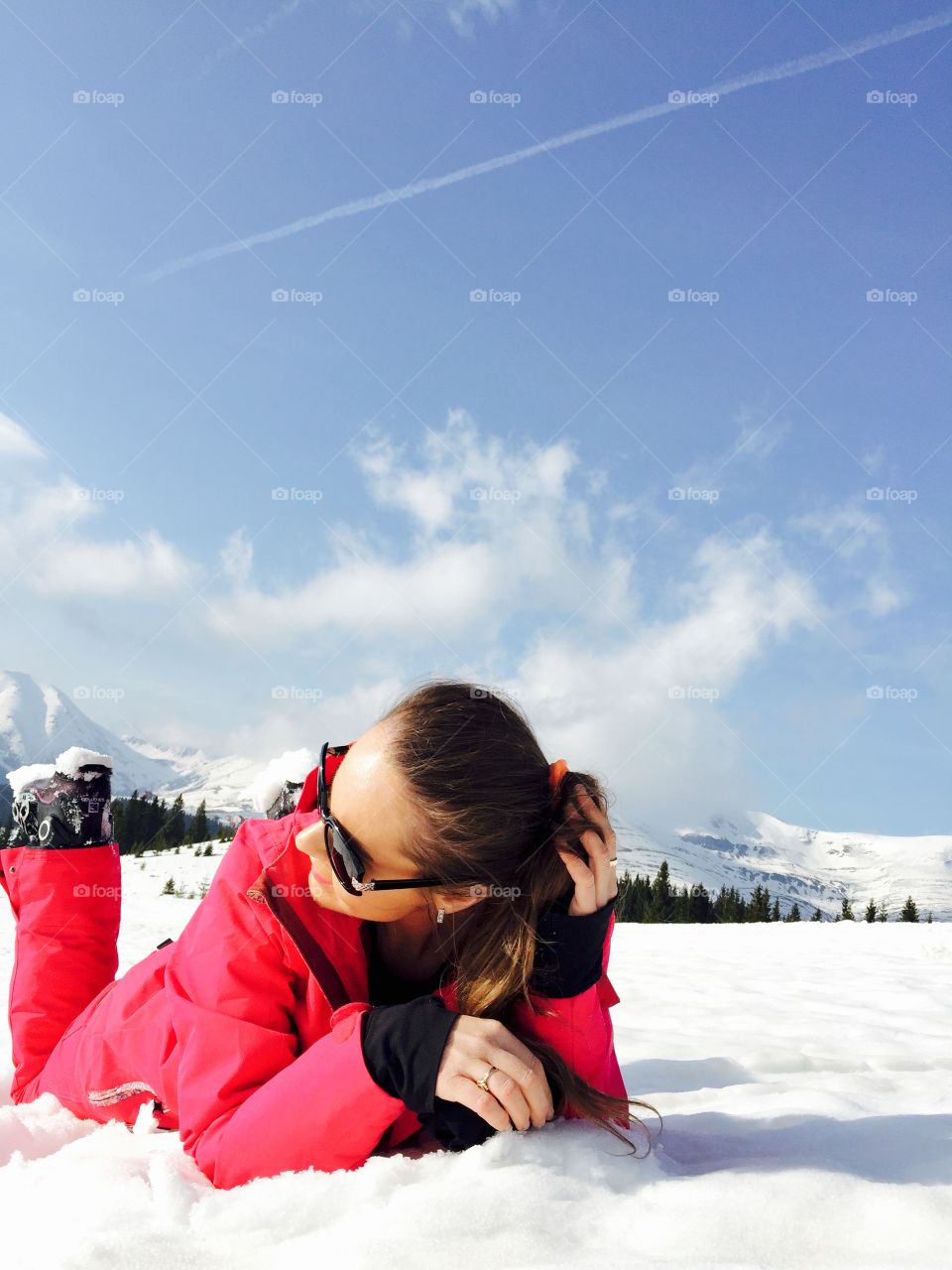 Smiling woman with sunglasses dressed in pink ski costume laying on snow with snow mountains in the background