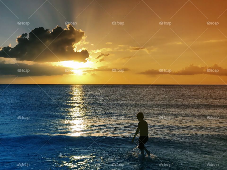 A fantastic golden sunset reflects on the ocean as a small boy plays in the waves.