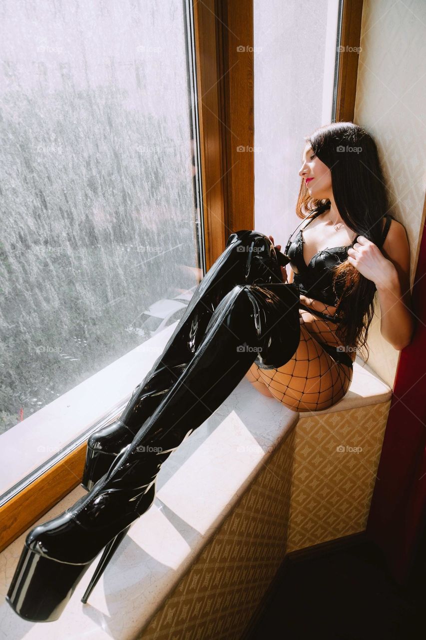 Girl in a black outfit, wearing long black knee boots with high heels, is sitting near a window, looking outside thoughtfully