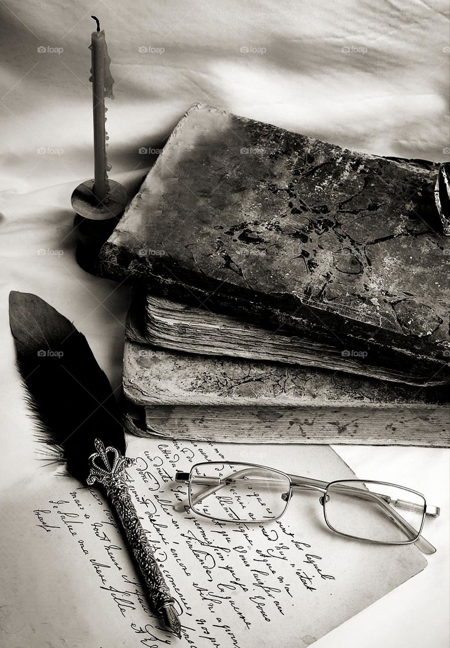 Black and white photo. Old books, page with text in French, glasses, extinguished candle, writing pen. Vintage items