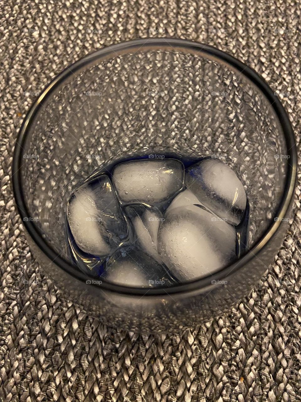 Ice cubes (aka frozen water) melting in a gray-tinted glass sitting on a woven gray placemat. 