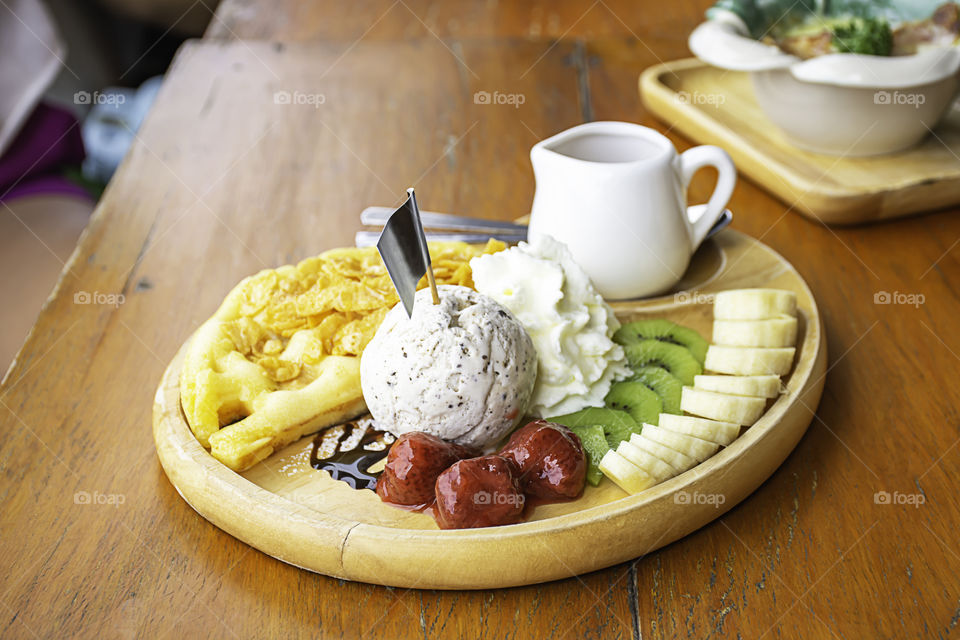 Sweet water pour on the waffle with ice cream and fruits including bananas, kiwi and strawberries in wooden plate on table.