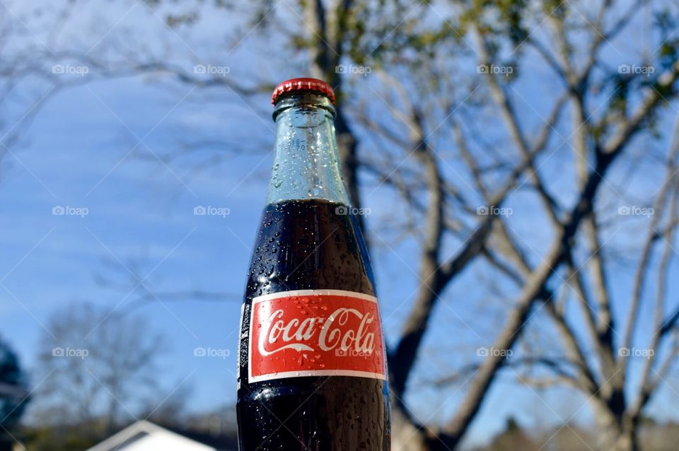 A glass bottle of Coca-Cola outside with A tree and a rooftop in the background