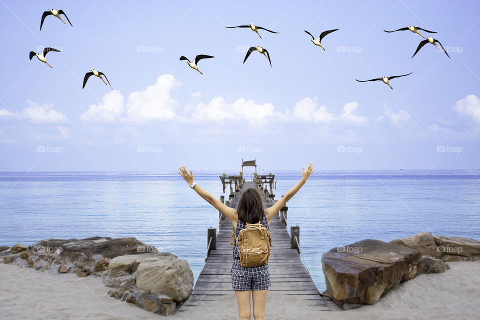 Women raise their arms and shoulder backpack on wooden bridge pier boat in the sea and the birds flying in the sky at Koh Kood, Trat in Thailand.