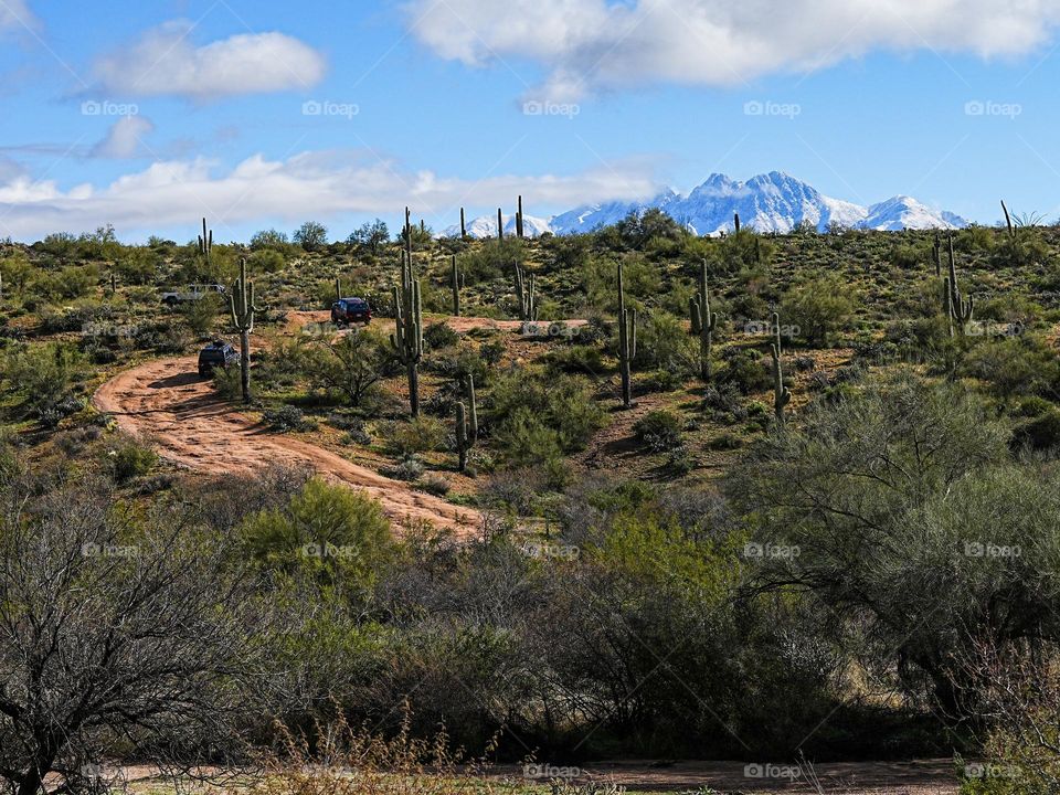 Four wheel drive vehicles scramble up a desert off road path while snow capped mountains loom in the background