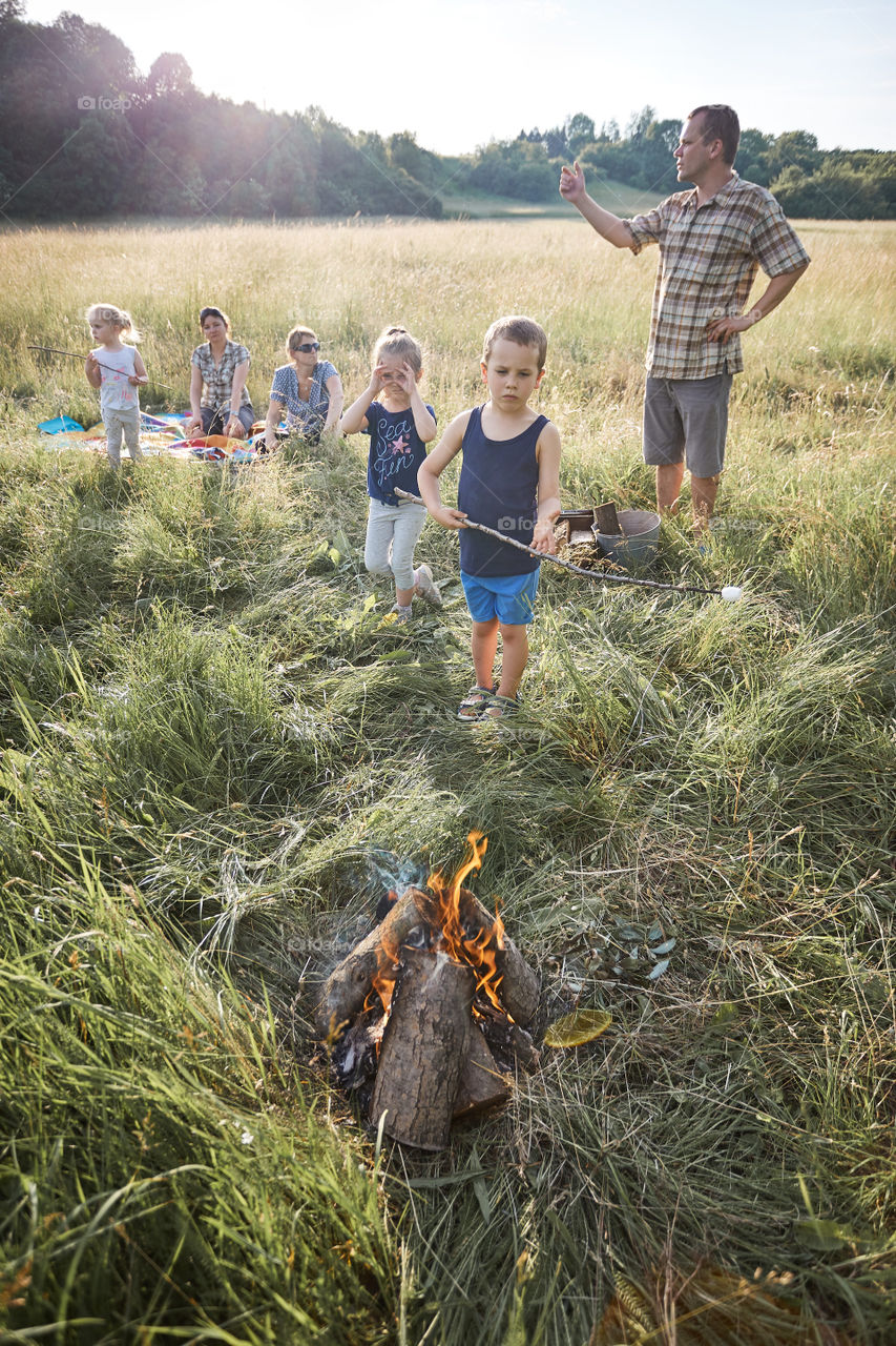 Little boy roasting marshmallow over a campfire. Family spending time together on a meadow, close to nature. Parents and children sitting on a blanket on grass. Candid people, real moments, authentic situations