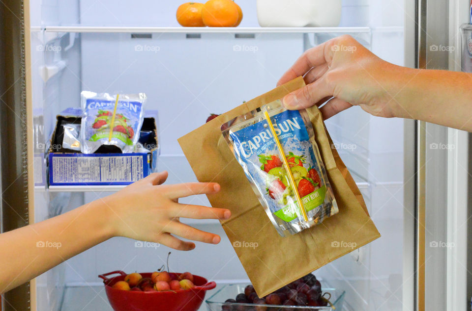 Parent handing child a bagged lunch with a drink pouch in front of an open refrigerator