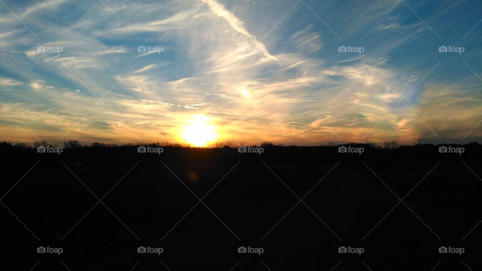 Stunning sunset. The ground is a shadowed, dark. The sun is bright and beginning to tuck beyond the horizon. Yellow and orange streak across a cloudy, blue sky.