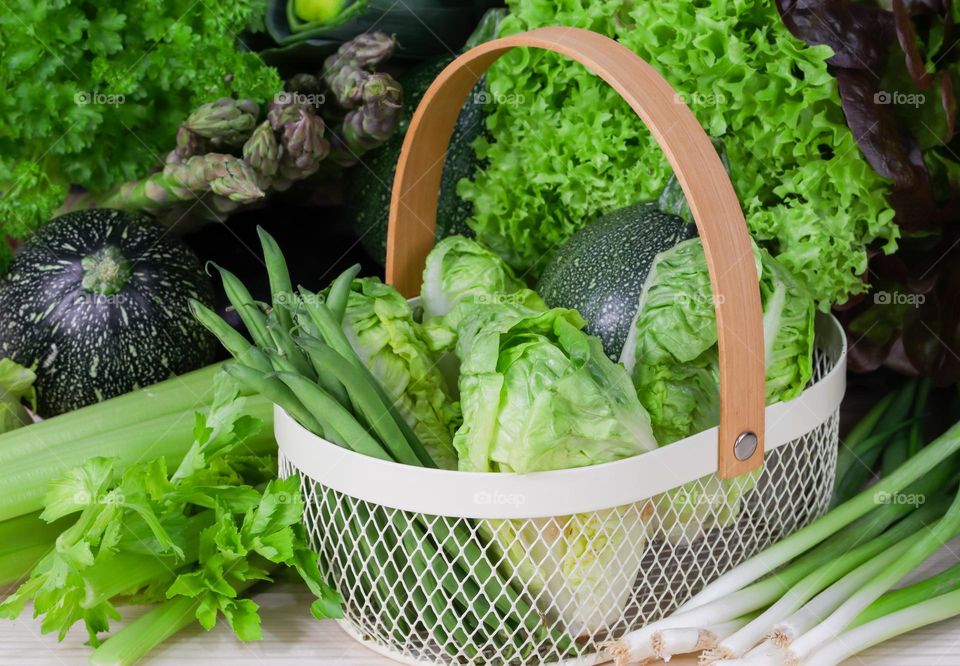 Green vegetables from green beans,round zucchini,curly lettuce in a wicker metal basket on a wooden table with green onions,celery and lettuce leaves around it. The concept of a basket of green vegetables and healthy eating.