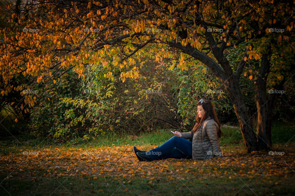 Girl in autumn sits under a tree and looks at the phone that she holds in her hand, everything is strewn with yellow leaves fallen from the tree.