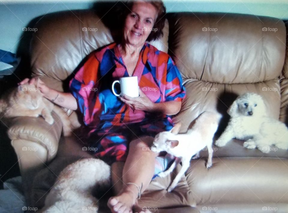 Woman in robe having coffee, sitting on couch with 4 pets, smiling.