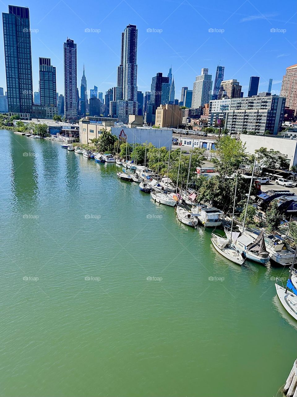 This is beautiful “Newtown Creek” with its many boats seen from the “Pulaski Bridge” that connects “Greenpoint”, Brooklyn to LIC, Queens. Across the “East River” we see “Manhattan” including the “Empire State Building”. 2024. Hypnotic Productions