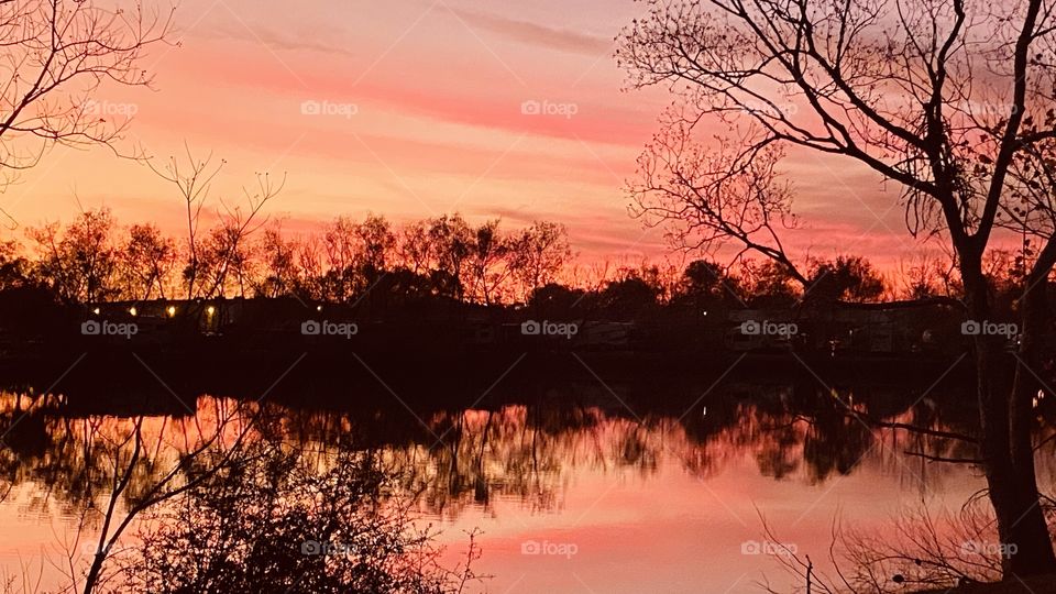 Twilight is in full view over reflections of Landscape of Lake Water. Clouds cover sky with Hues of Pink Yellow Some streaks of Purple.