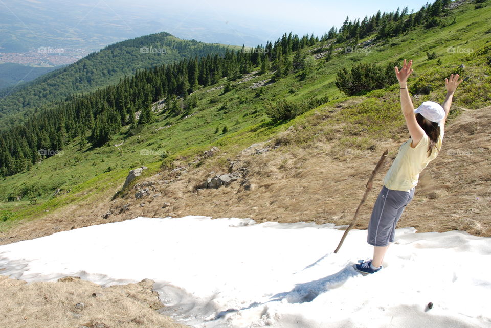 A path of white snow on a mountain slope in mid summer with green grass and trees around