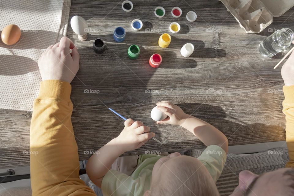 Dad teaches a small child to draw with bright, multi-colored gouache paints on a gray, wooden table